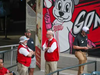 Four figures in black and red stand doing security detail in front of a Richmond Flying Squirrels image on a wall.